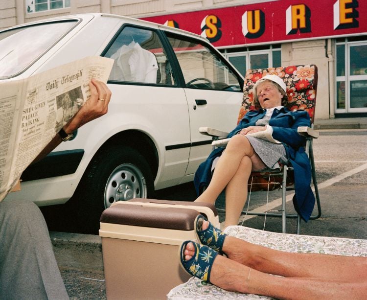 A woman sleeping in a camping chair on a parking lot next to a car